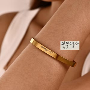 Pode incluir: Uma pulseira de metal dourado com a palavra always em letra cursiva, seguida de um pequeno símbolo de coração. O mesmo texto e design de coração também são exibidos em uma caixa retangular branca separada ao lado da pulseira.