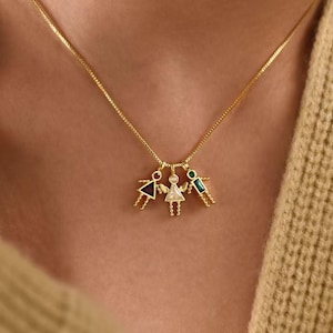 Puede incluir: Un collar dorado con tres colgantes. Cada colgante es una figura estilizada con detalles coloridos. El collar se lleva sobre un suéter beige. El diseño de la cadena es fino y delicado.