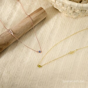 Puede incluir: Dos delicados collares. Uno es de oro rosa con una piedra preciosa azul y un dije de ala. El otro es dorado con una piedra preciosa verde y un dije de ala. Los collares se exhiben sobre una superficie beige claro.