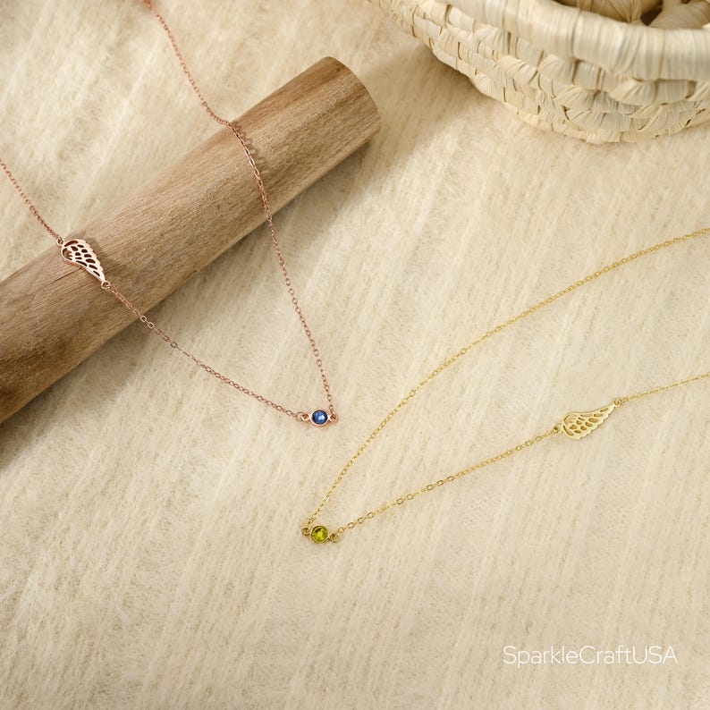 Puede incluir: Dos delicados collares. Uno es de oro rosa con una piedra preciosa azul y un dije de ala. El otro es dorado con una piedra preciosa verde y un dije de ala. Los collares se exhiben sobre una superficie beige claro.