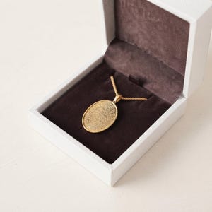 May include: A gold-toned oval pendant necklace featuring a detailed fingerprint engraving on the front surface. The pendant is attached to a matching gold-toned chain and is displayed inside a white jewelry box with a dark brown velvet interior.
