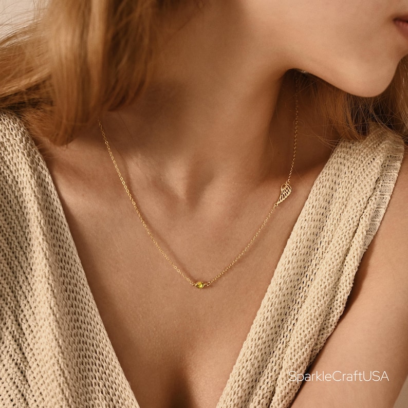 Puede incluir: Un delicado collar de oro con un peque&ntilde;o colgante de piedra preciosa amarilla y un dije de ala. El collar se lleva puesto y se muestra sobre un fondo neutro. La joyer&iacute;a es sencilla y elegante.