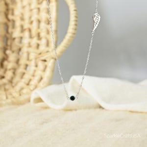 Puede incluir: Un delicado collar de plata con una peque&ntilde;a cuenta negra y un dije de ala. El collar se exhibe sobre una tela blanca suave, con una cesta tejida en el fondo. El collar es una pieza de joyer&iacute;a simple y elegante.