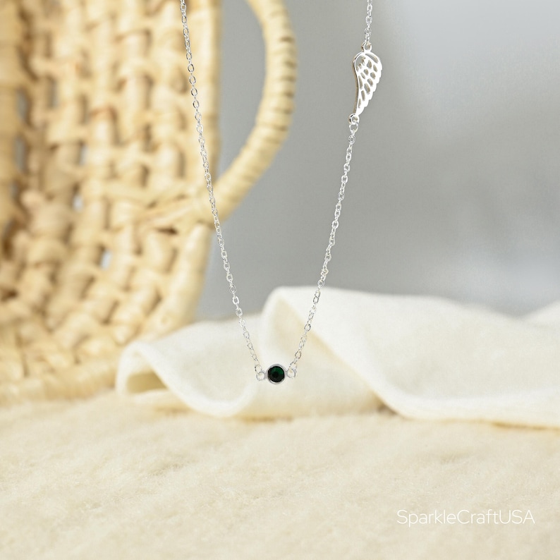 Puede incluir: Un delicado collar de plata con una peque&ntilde;a cuenta negra y un dije de ala. El collar se exhibe sobre una tela blanca suave, con una cesta tejida en el fondo. El collar es una pieza de joyer&iacute;a simple y elegante.