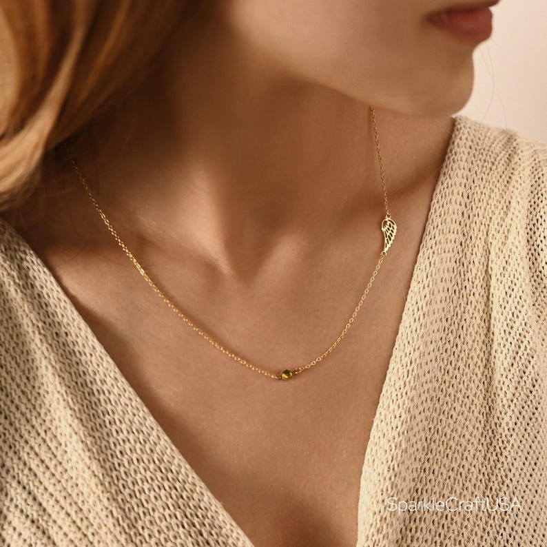 Puede incluir: Un delicado collar dorado con una peque&ntilde;a gema verde y un dije en forma de ala. El collar lo lleva una persona, con la cadena descansando sobre la clav&iacute;cula. El fondo es un su&eacute;ter color crema.
