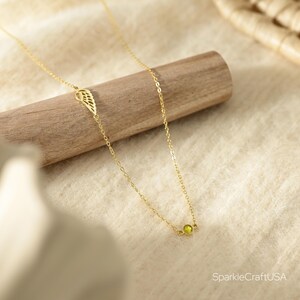 Puede incluir: Un delicado collar dorado con una peque&ntilde;a gema redonda de color verde claro y un dije en forma de ala. El collar se exhibe sobre una clavija de madera sobre un fondo de tela neutra.