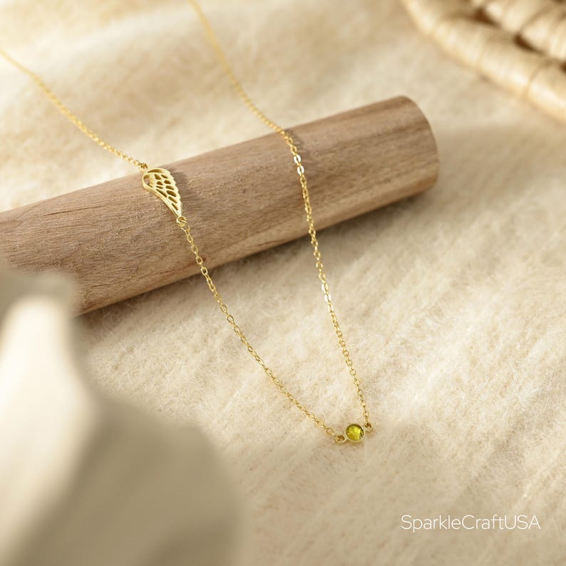 Puede incluir: Un delicado collar dorado con una peque&ntilde;a gema redonda de color verde claro y un dije en forma de ala. El collar se exhibe sobre una clavija de madera sobre un fondo de tela neutra.