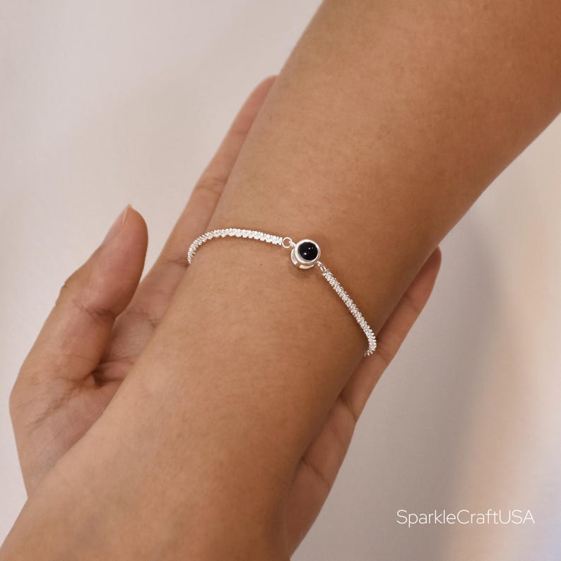 May include: A delicate silver bracelet with a small, round, black gemstone at the center. The bracelet is worn on a wrist, showcasing its simple yet elegant design. The bracelet's band is adorned with small, sparkling details.