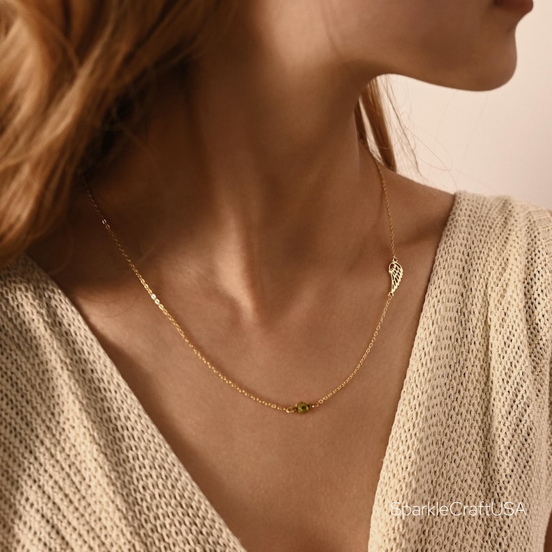 Puede incluir: Un delicado collar dorado con una peque&ntilde;a piedra verde y un colgante de ala estilizada. El collar lo lleva una persona, y el dise&ntilde;o es sencillo y elegante. El collar es una joya.
