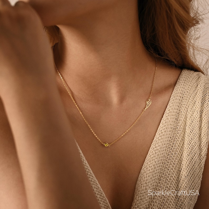 Puede incluir: Un delicado collar dorado con un peque&ntilde;o colgante de ala y una sola gema amarilla. El collar se lleva en el cuello de una persona, con la cadena apoyada en su piel. Una joya sencilla pero elegante.