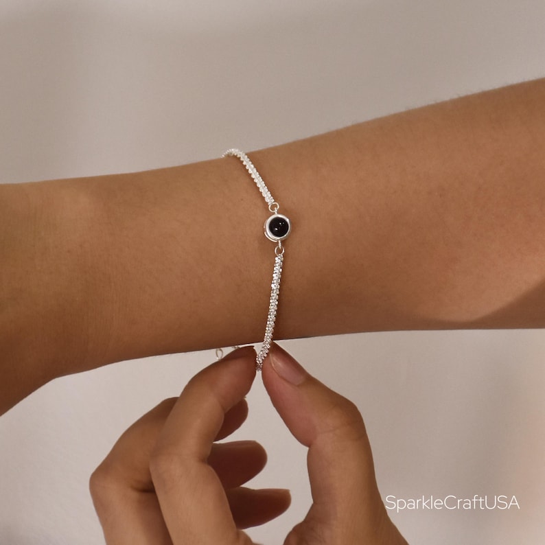 May include: A silver bracelet with a black gemstone and small, clear stones. The bracelet is being held by a person's hand. The bracelet is delicate and elegant.