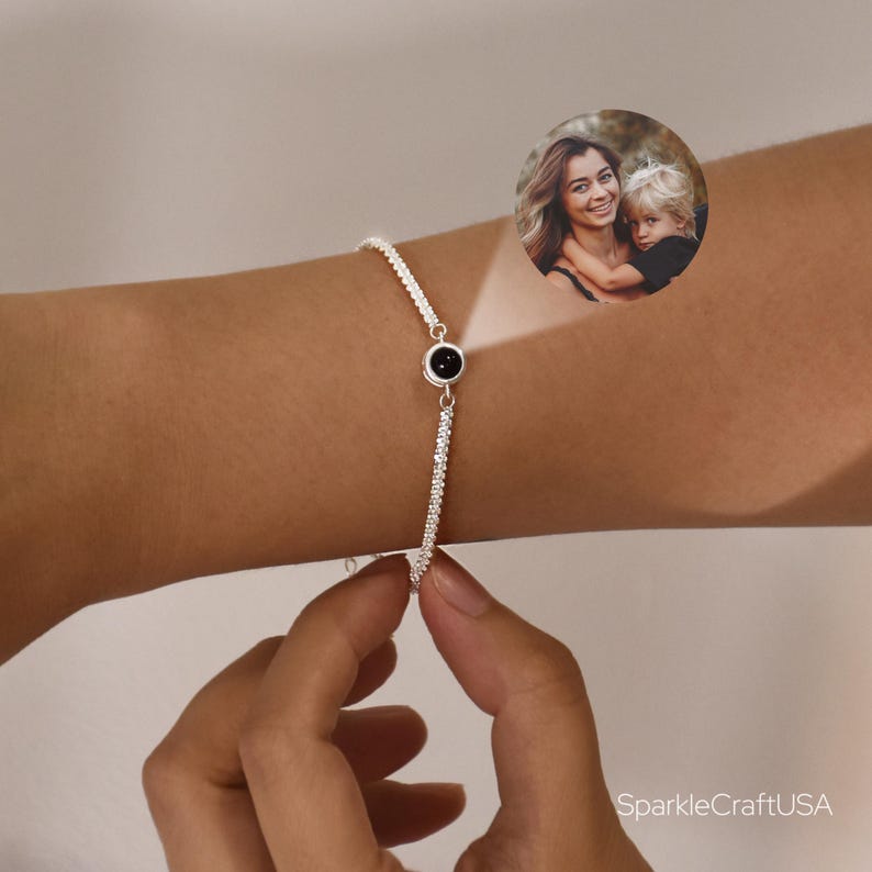 May include: A silver bracelet with a black circular stone in the center. The bracelet is adorned with small, sparkling stones. The image includes a small photo of a woman and child. The bracelet is being held by a hand.