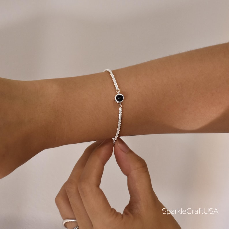 May include: A silver bracelet with a black circular stone in the center. The bracelet is made of small, sparkling beads. The bracelet is being worn on a person's wrist.