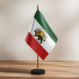 Puede incluir: Una pequeña bandera de mesa con la bandera iraní. La bandera tiene una franja roja en la parte inferior, una franja blanca en el medio y una franja verde en la parte superior. El centro de la bandera muestra un león dorado sosteniendo una espada frente a un sol.