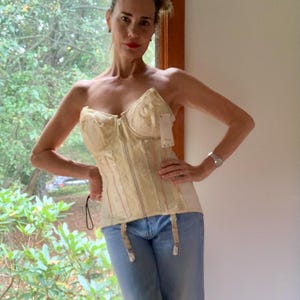 May include: Cream-colored corset with a front zipper and attached garter straps. The corset is paired with light blue jeans. A tag is attached to the corset. The background includes a window with greenery.