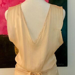 May include: A sleeveless, light peach-colored dress with a deep V-neckline and a matching tie belt. The dress appears to be made of a soft, flowing fabric. The back of the dress is visible, showcasing the design.