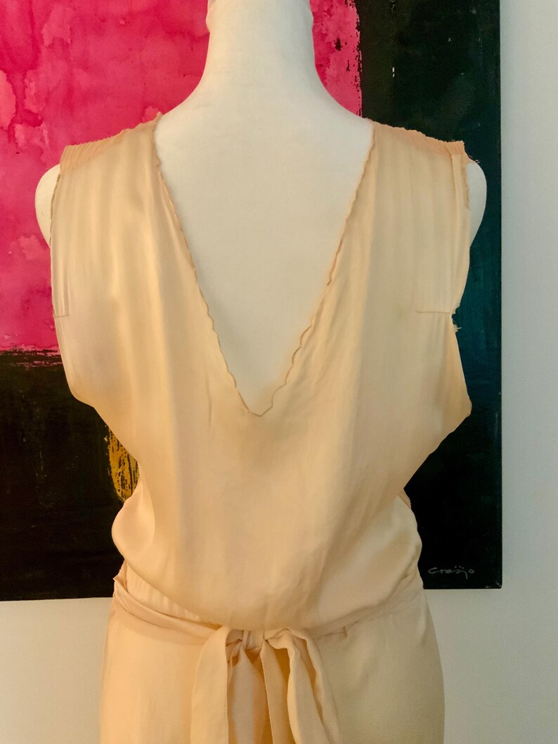 May include: A sleeveless, light peach-colored dress with a deep V-neckline and a matching tie belt. The dress appears to be made of a soft, flowing fabric. The back of the dress is visible, showcasing the design.