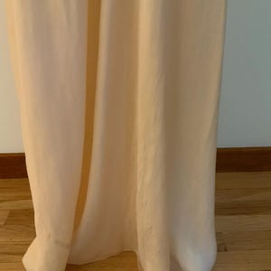 May include: A floor-length, pale peach-colored dress made of a flowing, lightweight fabric. The dress has a simple, elegant design, with a subtle sheen. The hemline gently pools on the wooden floor.