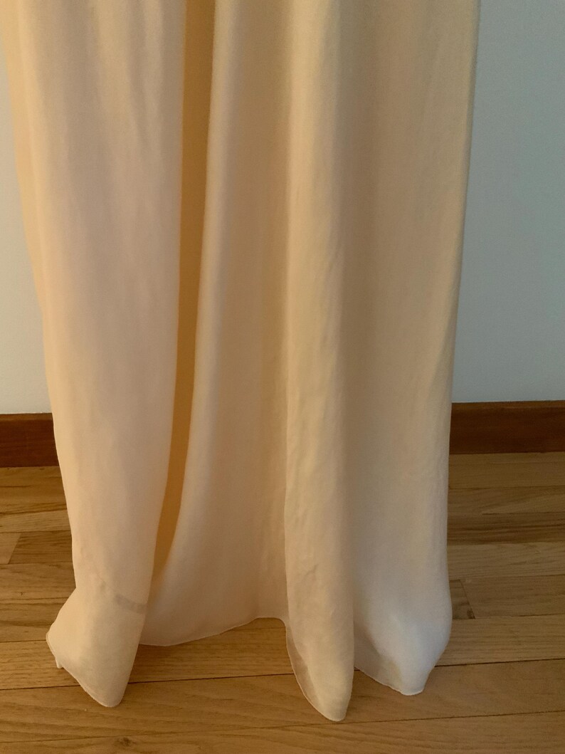 May include: A floor-length, pale peach-colored dress made of a flowing, lightweight fabric. The dress has a simple, elegant design, with a subtle sheen. The hemline gently pools on the wooden floor.