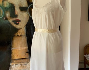 Puede incluir: Un camisón blanco de estilo vintage sobre un maniquí. El camisón presenta un escote con volantes, detalles bordados y un cinturón de cinta de satén. El vestido, a la altura de la rodilla, tiene un diseño clásico y elegante. El fondo incluye una pintura y un suelo de madera.