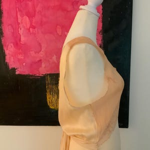 May include: A peach-colored satin slip dress with a delicate, pleated chiffon overlay on the bodice. The dress is displayed on a mannequin, with a bow detail at the back. The background features a pink and black abstract painting with the text "fuerza natural".