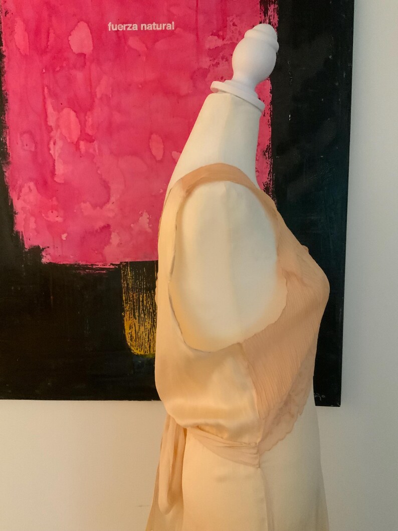 May include: A peach-colored satin slip dress with a delicate, pleated chiffon overlay on the bodice. The dress is displayed on a mannequin, with a bow detail at the back. The background features a pink and black abstract painting with the text "fuerza natural".