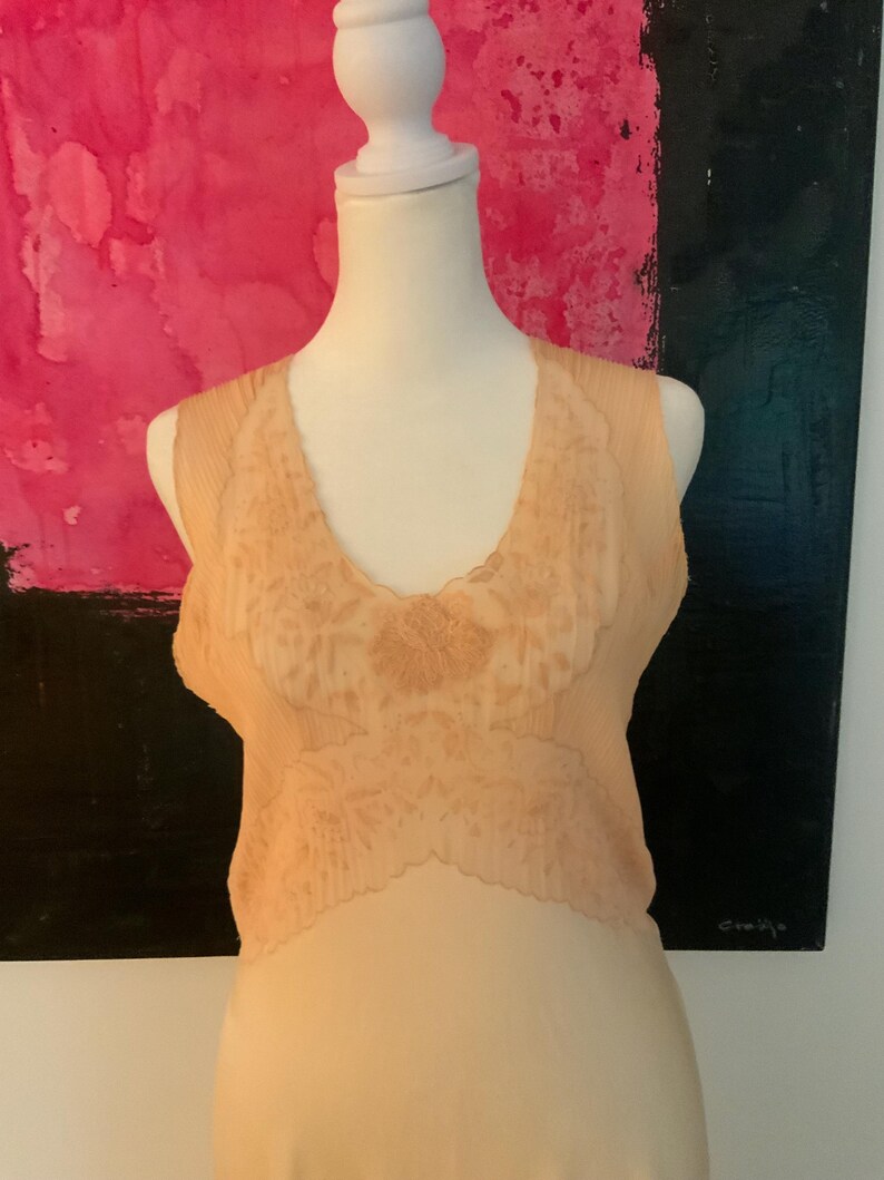 May include: A peach-colored vintage slip dress with a delicate lace bodice. The slip is displayed on a mannequin, showcasing its soft, flowing fabric and intricate floral lace detailing. The dress has a scoop neckline and a subtle sheen.