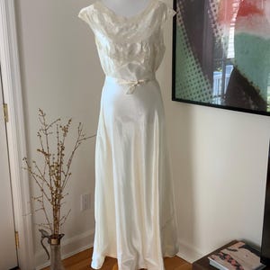 Pode incluir: Um vestido branco de cetim com um corpete de renda e uma faixa na cintura. O vestido est&aacute; em um manequim e &eacute; exibido em uma sala com piso de madeira.