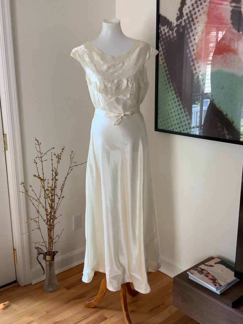 Pode incluir: Um vestido branco de cetim com um corpete de renda e uma faixa na cintura. O vestido est&aacute; em um manequim e &eacute; exibido em uma sala com piso de madeira.
