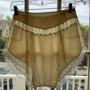 Könnte beinhalten: Vintage High-Waist-Unterwäsche aus transparentem, goldgelbem Stoff. Die Kanten sind mit zarter weißer Spitze verziert. Das Kleidungsstück hängt an einem transparenten Kunststoffbügel vor einem Fenster.
