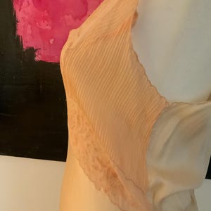 May include: A peach-colored slip dress with a pleated bodice and lace trim. The dress has a flowing, silky appearance and a delicate, feminine design. The dress is displayed on a mannequin.