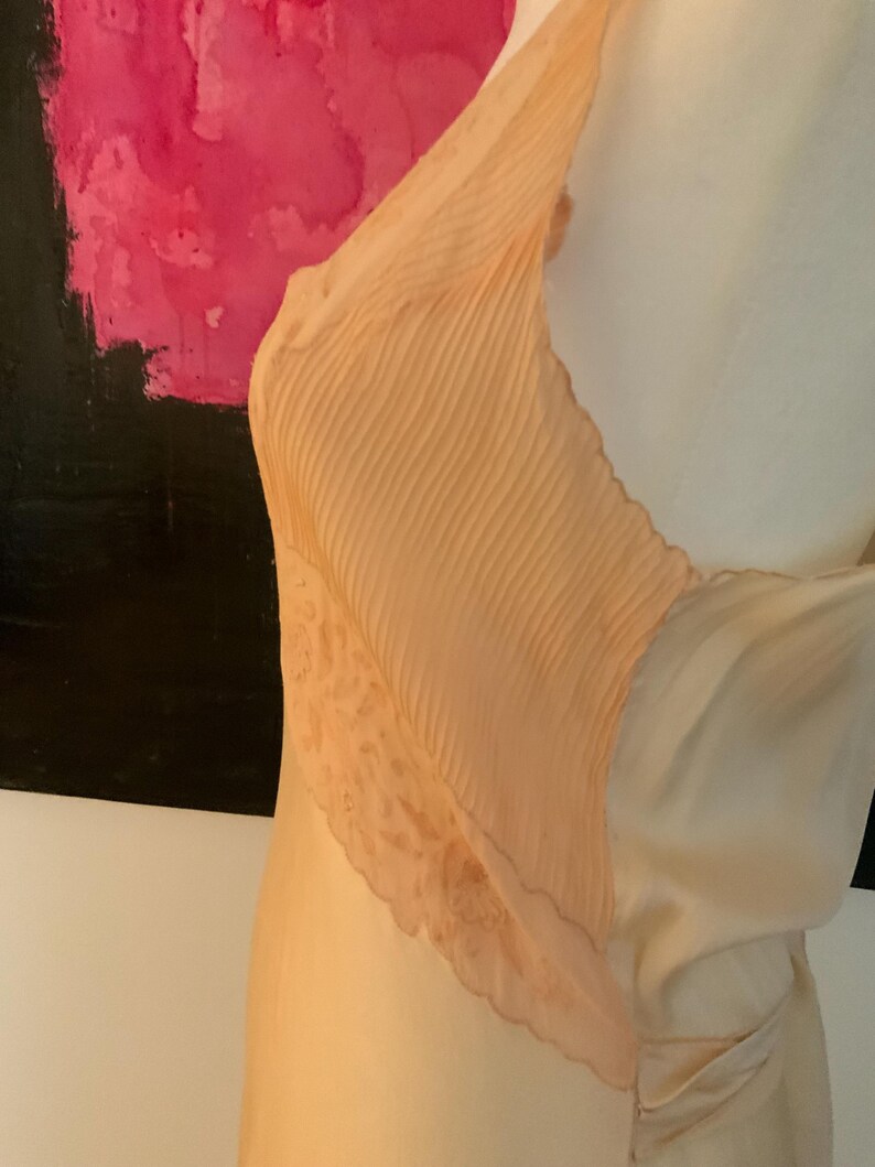 May include: A peach-colored slip dress with a pleated bodice and lace trim. The dress has a flowing, silky appearance and a delicate, feminine design. The dress is displayed on a mannequin.