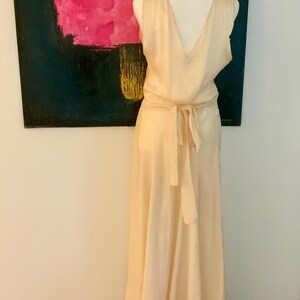 May include: A sleeveless, pale peach-colored dress with a V-neck and a tie-waist. The dress is displayed on a mannequin. The background features a painting with pink and black colors and the text "fuerza natural".