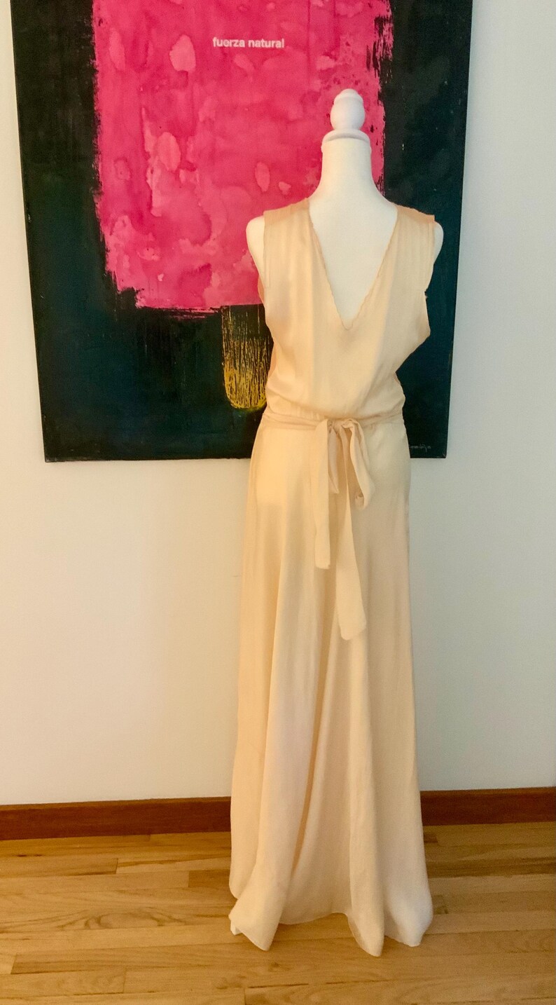 May include: A sleeveless, pale peach-colored dress with a V-neck and a tie-waist. The dress is displayed on a mannequin. The background features a painting with pink and black colors and the text "fuerza natural".