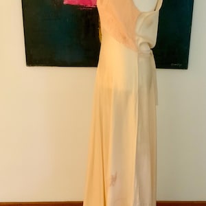 May include: A vintage-style, floor-length gown in a soft, creamy peach color. The bodice features a delicate lace detail, and the dress has a flowing, elegant silhouette. The dress is displayed on a mannequin, showcasing its design and drape.