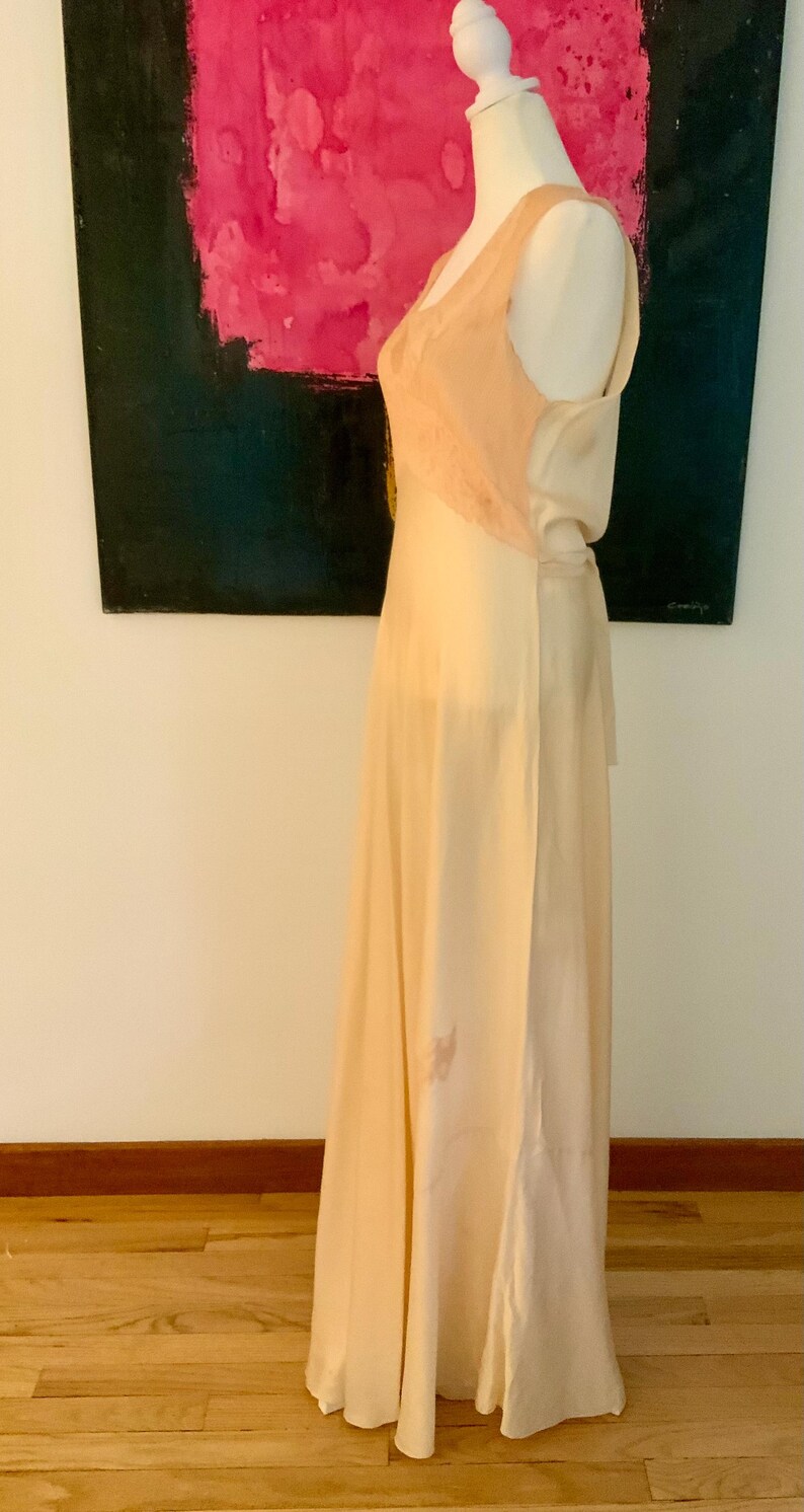 May include: A vintage-style, floor-length gown in a soft, creamy peach color. The bodice features a delicate lace detail, and the dress has a flowing, elegant silhouette. The dress is displayed on a mannequin, showcasing its design and drape.