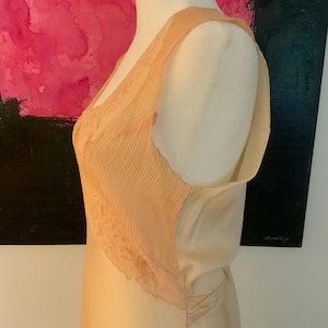 May include: A peach-colored slip dress with a pleated bodice and lace trim. The dress is displayed on a mannequin, showcasing its delicate fabric and design. The dress has a V-neckline and a flowing skirt, suggesting a vintage or romantic style.