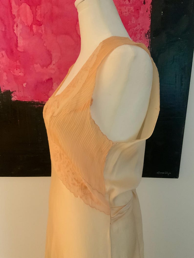 May include: A peach-colored slip dress with a pleated bodice and lace trim. The dress is displayed on a mannequin, showcasing its delicate fabric and design. The dress has a V-neckline and a flowing skirt, suggesting a vintage or romantic style.