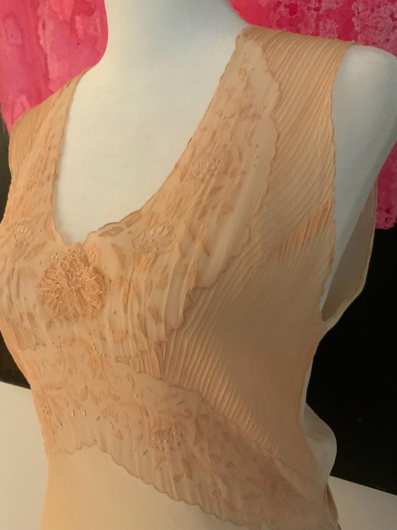 May include: A peach-colored, sleeveless dress with a V-neckline. The dress features delicate floral embroidery and pleated fabric details. The dress is displayed on a mannequin.