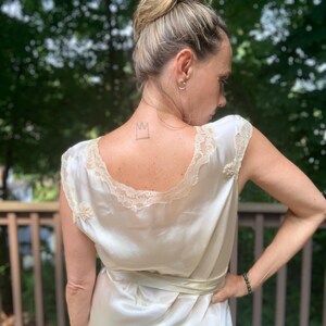 Pode incluir: Um vestido de camisola de cetim branco com uma borda de renda e um la&ccedil;o de fita. O vestido tem uma coroa desenhada nas costas.