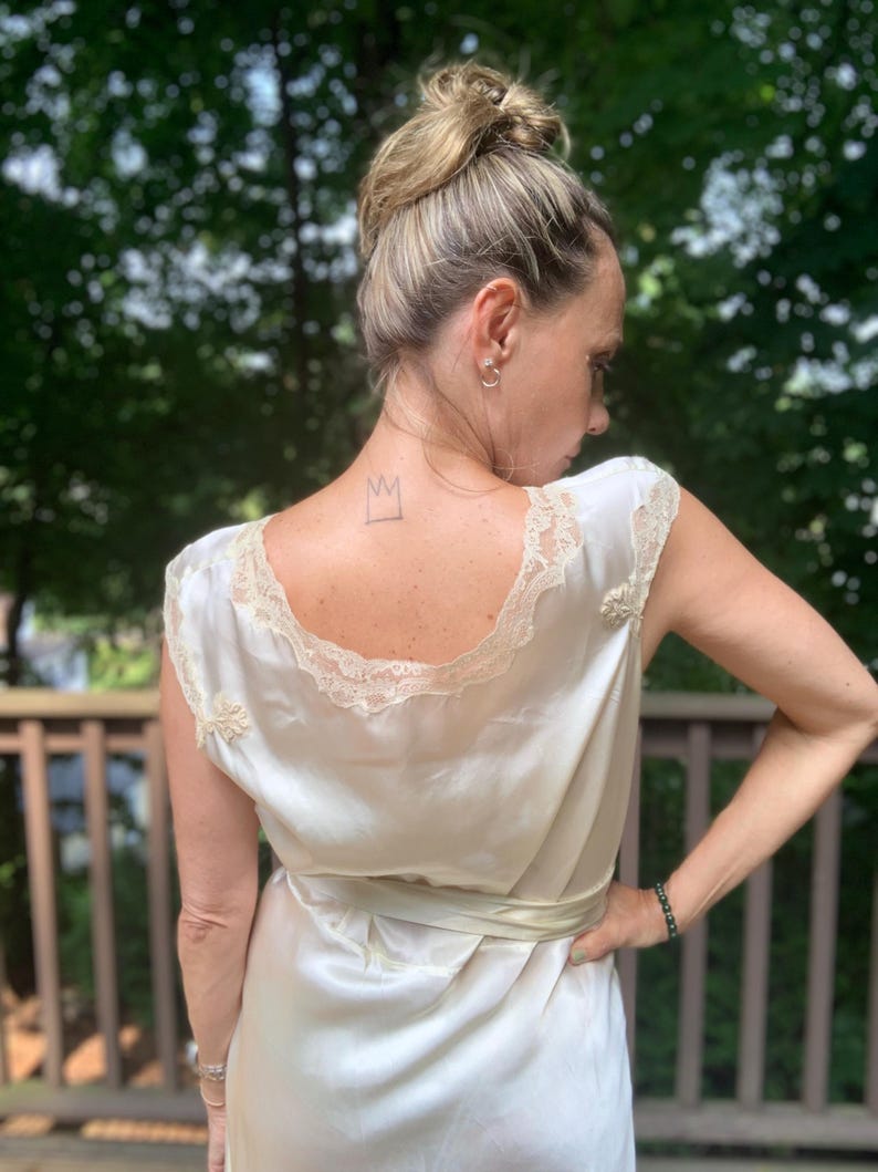 Pode incluir: Um vestido de camisola de cetim branco com uma borda de renda e um la&ccedil;o de fita. O vestido tem uma coroa desenhada nas costas.