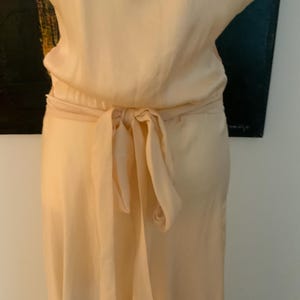 May include: A light peach-colored dress with a tie-waist detail. The dress has a flowing skirt and a matching tie belt that is knotted in the back. The fabric appears to be a soft, lightweight material.