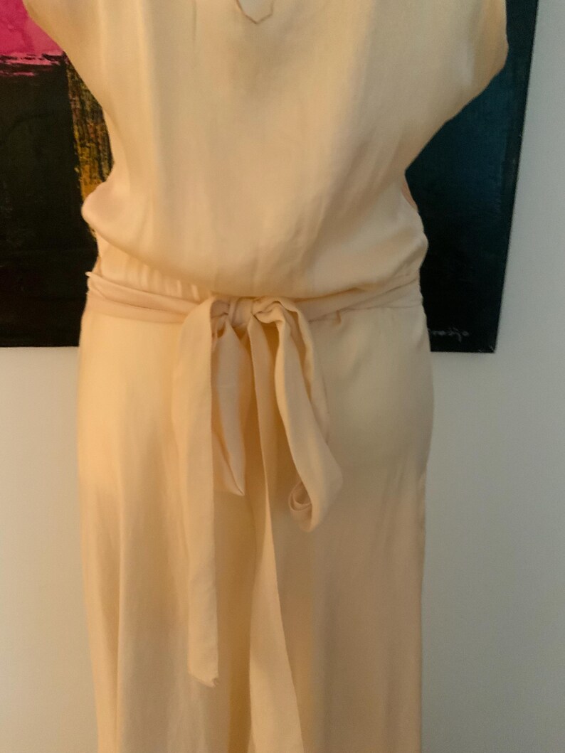 May include: A light peach-colored dress with a tie-waist detail. The dress has a flowing skirt and a matching tie belt that is knotted in the back. The fabric appears to be a soft, lightweight material.