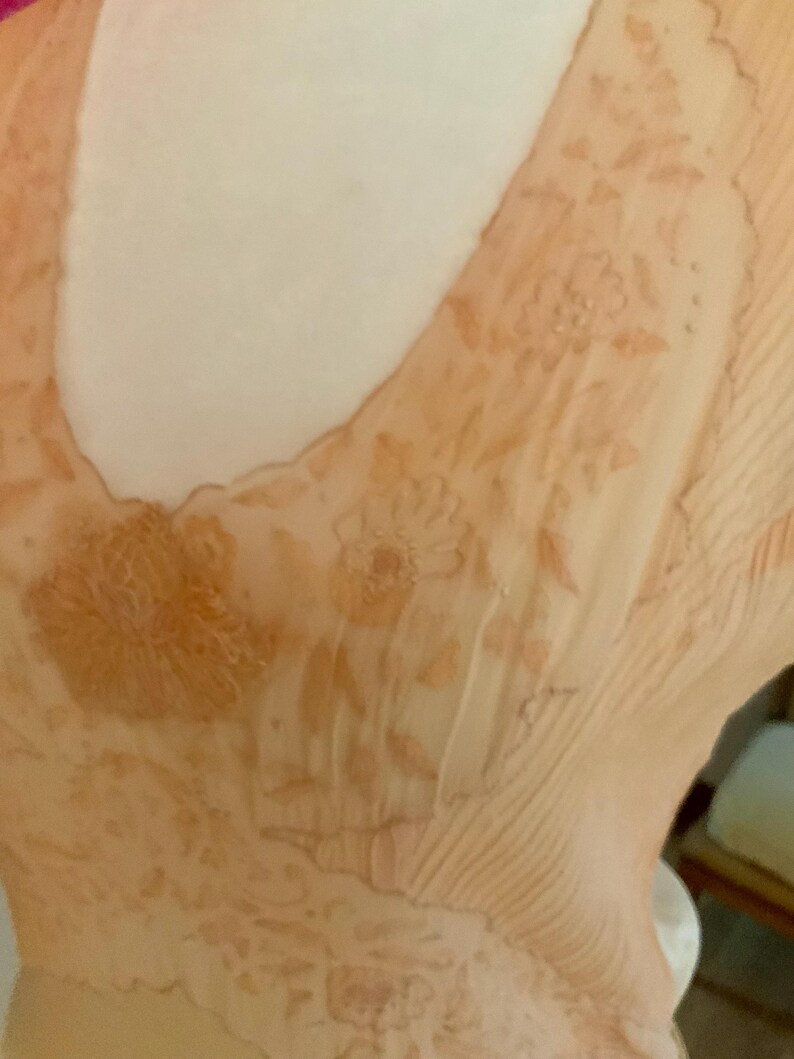 May include: Close-up of a peach-colored garment with a V-neckline. The fabric is semi-sheer and adorned with floral patterns in a darker peach hue. The garment has a delicate, flowing appearance, suggesting a vintage or romantic style.