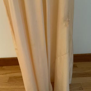 May include: A floor-length, pale peach-colored dress with soft folds. The fabric drapes gracefully, creating a flowing silhouette. The dress appears to be made of a lightweight material, suitable for warmer weather. A small embellishment is visible near the hem.
