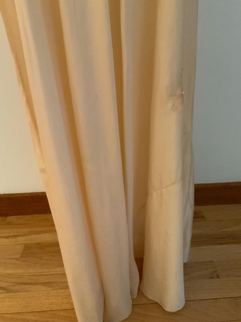 May include: A floor-length, pale peach-colored dress with soft folds. The fabric drapes gracefully, creating a flowing silhouette. The dress appears to be made of a lightweight material, suitable for warmer weather. A small embellishment is visible near the hem.
