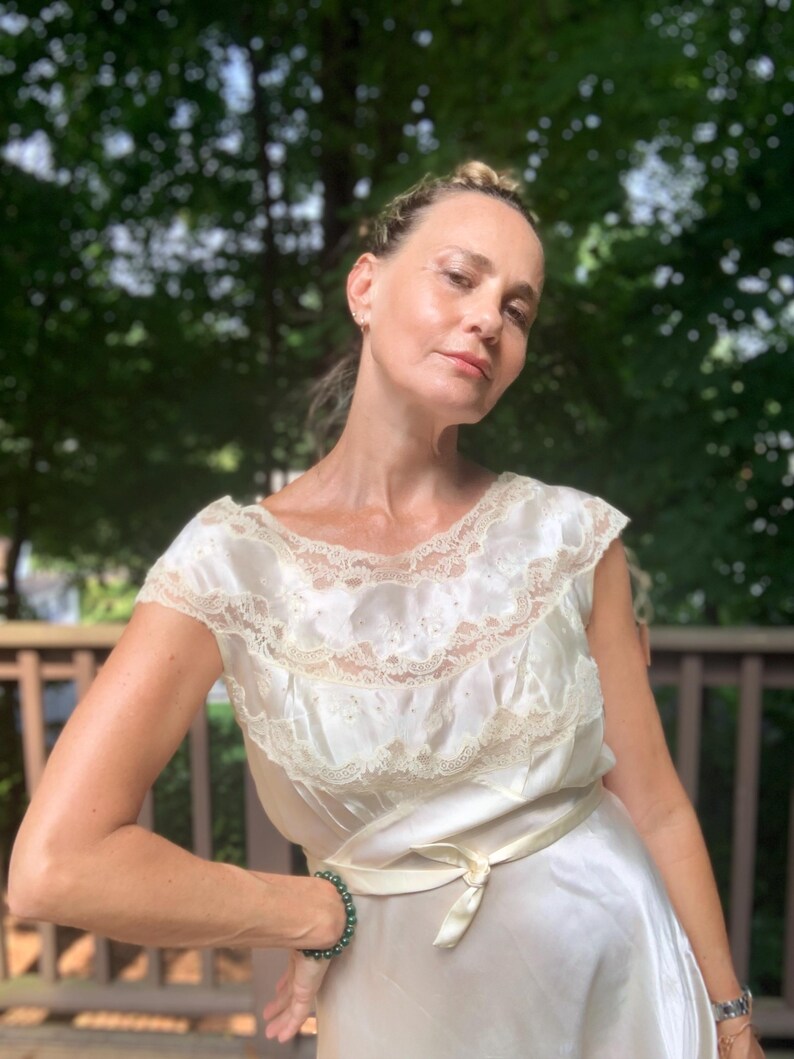 Pode incluir: Uma mulher vestindo um vestido de camisola de cetim branco com uma borda de renda e uma faixa de cetim. Ela est&aacute; usando uma pulseira de contas verdes no pulso.