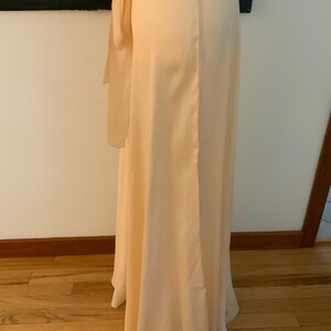 May include: A floor-length, pale peach-colored dress with a flowing silhouette. The dress features a wrap-style bodice and a long, elegant skirt. A matching sash is tied at the waist, adding a touch of detail. The fabric appears to be a soft, silky material.