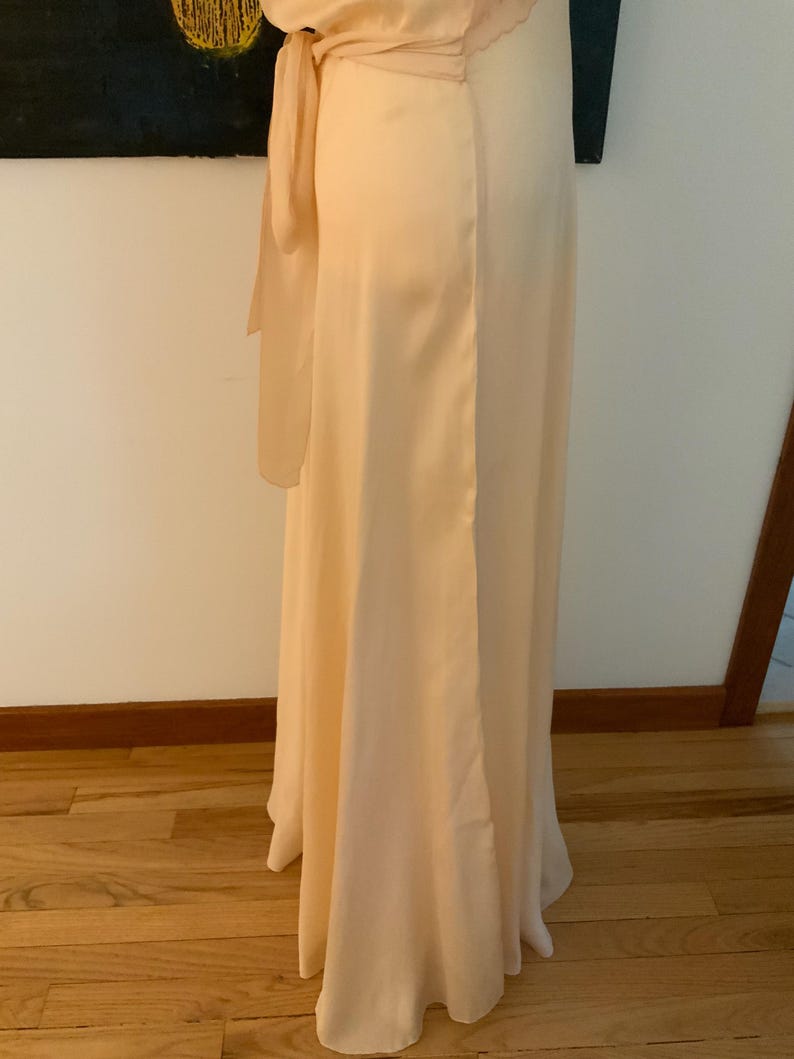 May include: A floor-length, pale peach-colored dress with a flowing silhouette. The dress features a wrap-style bodice and a long, elegant skirt. A matching sash is tied at the waist, adding a touch of detail. The fabric appears to be a soft, silky material.