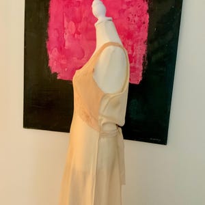 May include: A cream-colored silk dress on a mannequin. The dress features a lace panel on the bodice and a tie at the waist. The background includes a painting with the words "fuerza natural".